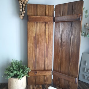 May include: Two wooden shutters with a dark brown stain, each with a horizontal wooden brace. The shutters are standing upright on a wooden surface.