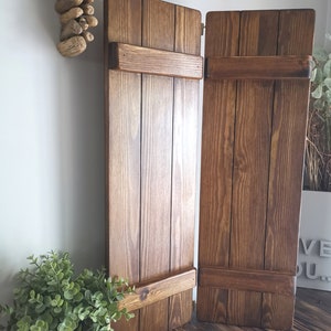 May include: Two brown wooden shutters with a rustic finish. The shutters are closed and have a simple design with horizontal slats and a single hinge.