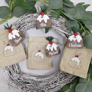 May include: Four Christmas-themed gift bags with personalized wooden ornaments. Each ornament is shaped like a Christmas pudding and features a different name: Alex, Brian, Christina, and Michael.