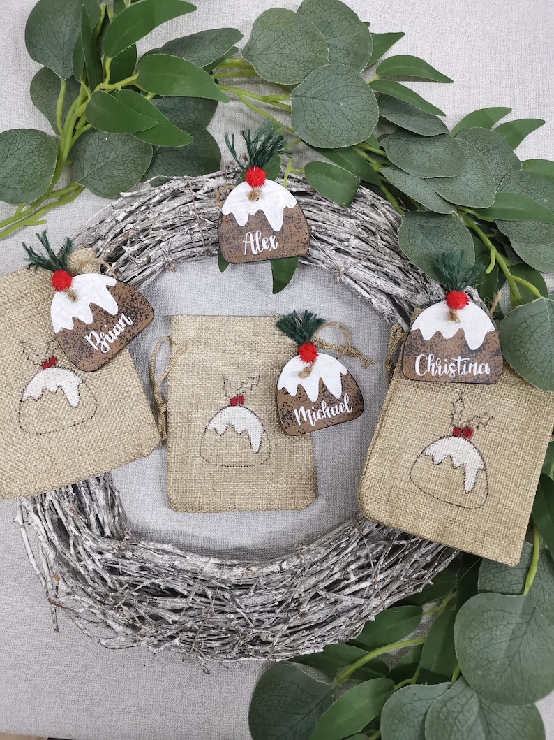 May include: Four Christmas-themed gift bags with personalized wooden ornaments. Each ornament is shaped like a Christmas pudding and features a different name: Alex, Brian, Christina, and Michael.