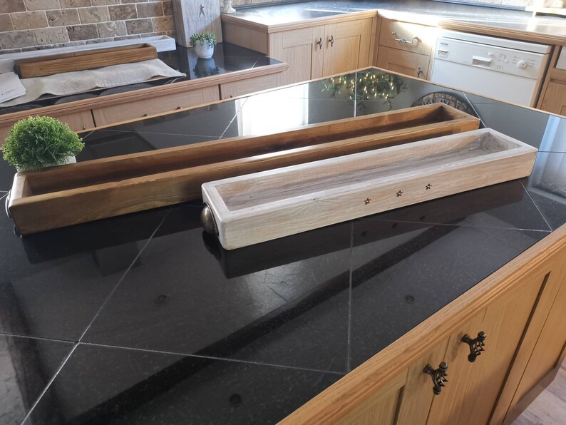 Op de afbeelding: Twee houten trays, een van naturel hout en een witgeschilderd, met een sterrendesign. De trays staan op een zwart aanrecht.