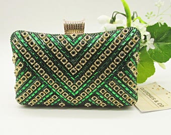 Puede incluir: Clutch rectangular verde con cuentas y cierre dorado. El clutch presenta un patrón geométrico de cuentas negras y doradas. El diseño se acentúa con adornos negros y dorados. Un accesorio elegante para ocasiones especiales.