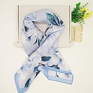 Op de afbeelding: Een lichtblauwe sjaal met witte bloemen en groene bladeren. De sjaal is opgevouwen en op een witte doos geplaatst. De sjaal heeft een lichtblauwe rand. Een kaart met de tekst "CLUTCHES & CO" is zichtbaar.