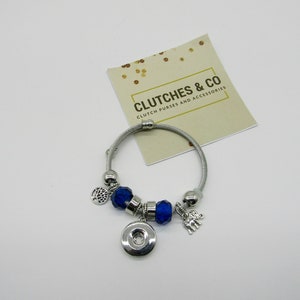 Puede incluir: Un brazalete de plata con cuentas de cristal azul y dijes de plata, incluyendo un árbol, un elefante y un botón. El brazalete está sobre un fondo blanco con una tarjeta que dice "Clutches & Co. Clutch purses and accessories."