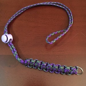 May include: A purple and green camouflage lanyard with a white plastic bead and a silver metal ring.