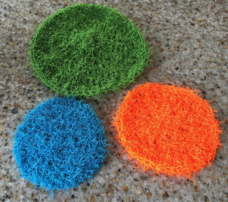 May include: Three round, crocheted scrubbers in green, blue, and orange. The scrubbers are made of a soft, fluffy material and are perfect for cleaning dishes or other surfaces.