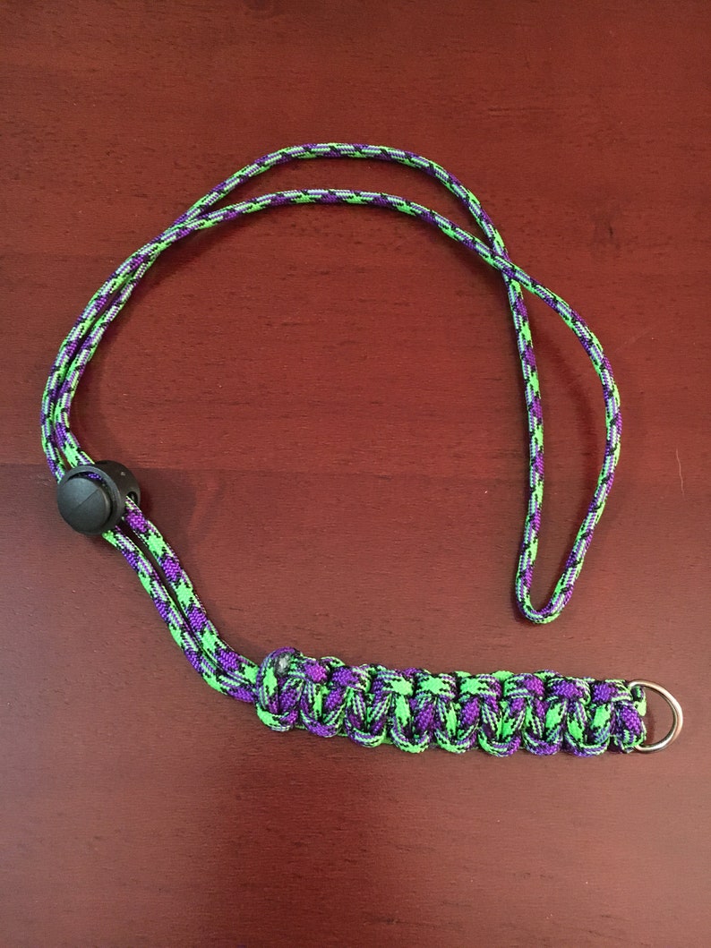 May include: A green and purple lanyard with a black plastic clip and a silver metal ring. The lanyard is made of braided cord.