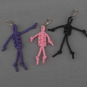 Puede incluir: Tres llaveros de paracord de colores en forma de figuras de palo. Los llaveros son morado, rosa y negro.