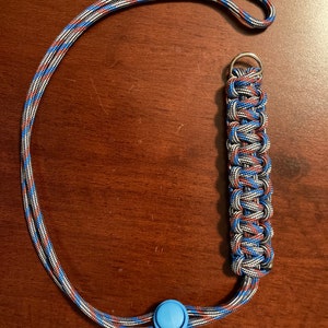 May include: A blue, red, and gray lanyard with a blue plastic button and a silver metal ring. The lanyard is made of braided cord.