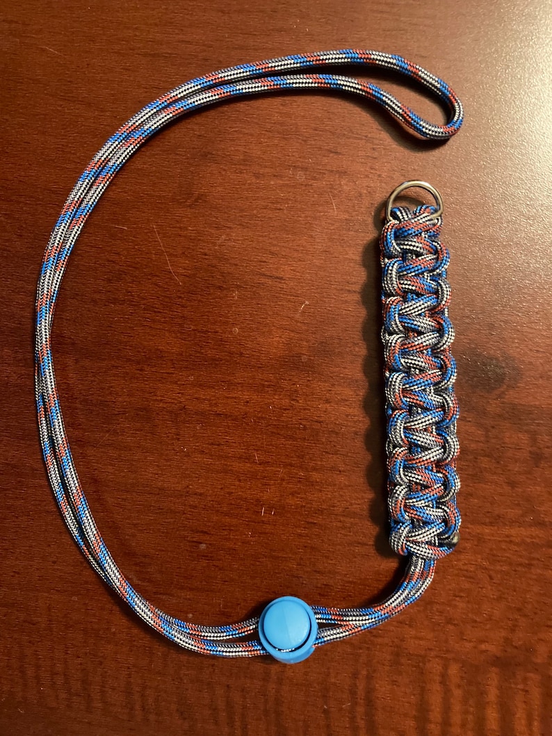 May include: A blue, red, and gray lanyard with a blue plastic button and a silver metal ring. The lanyard is made of braided cord.
