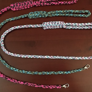 Puede incluir: Cuerdas de paracord trenzadas en varios colores, incluyendo rosa, azul y verde. Cada cuerda tiene un lazo en un extremo y un cierre metálico en el otro. Las cuerdas se muestran sobre una superficie marrón oscuro.
