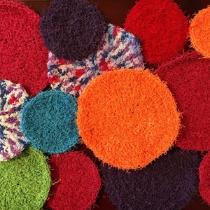 May include: A collection of colorful crocheted scrubbies in various colors, including red, orange, green, blue, and purple. The scrubbies are round and have a fuzzy texture.