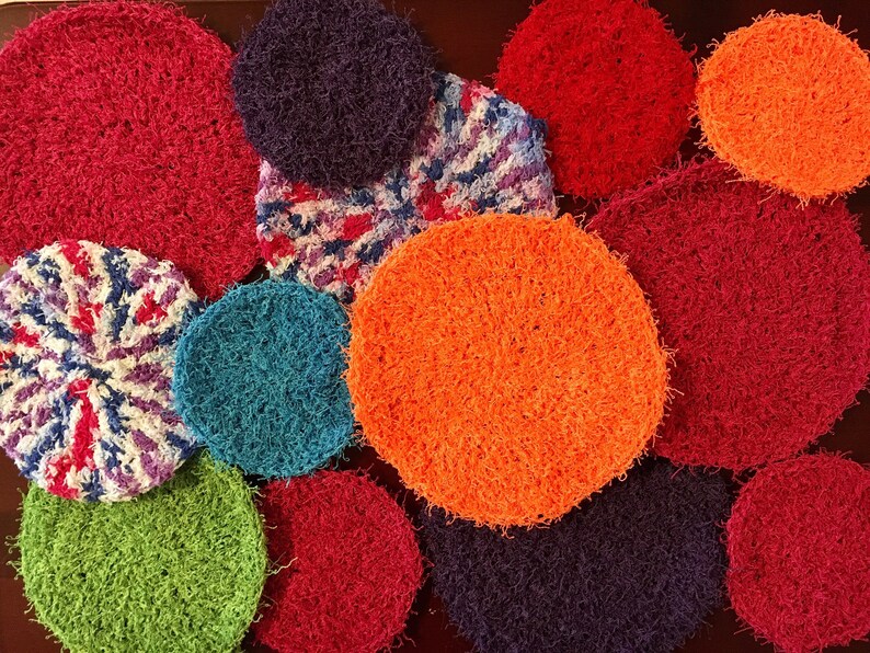 May include: A collection of colorful crocheted scrubbies in various colors, including red, orange, green, blue, and purple. The scrubbies are round and have a fuzzy texture.
