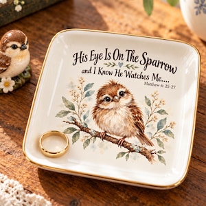 May include: A square white ceramic trinket dish with a gold-toned rim featuring a brown sparrow illustration perched on a branch, the text His Eye Is On The Sparrow and I Know He Watches Me.... Matthew 6: 25-27, and a plain gold-toned metal ring.