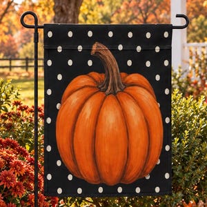 Peut inclure: Un drapeau de jardin rectangulaire présentant une grande citrouille orange centrale avec une tige brune sur un fond noir orné de petits pois blancs. Le drapeau est suspendu à un support en métal noir dans un jardin aux feuillages d'automne.