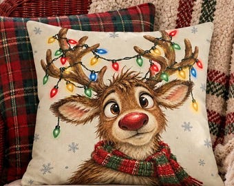 May include: A square throw pillow featuring a cartoon reindeer with a red nose, wearing a plaid scarf, and having colorful Christmas lights tangled in its antlers. The pillow is set against a cream background with small blue snowflake patterns.