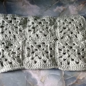 Op de afbeelding: Een handgemaakt, lichtgrijs gehaakt item, mogelijk een deken of kussensloop, bestaande uit meerdere vierkante granny squares die aan elkaar zijn gezet. Het garen heeft een zachte, getextureerde uitstraling. Het item wordt gepresenteerd op een gemarmerd oppervlak.