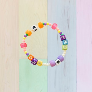 May include: A colorful beaded bracelet with the letters "OWA" spelled out in purple, blue, and green beads. The bracelet also features small orange and pink beads, as well as black heart charms.