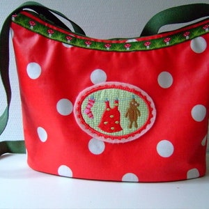 May include: Red polka dot fabric tote bag with a green strap and a white embroidered patch featuring a dress and a bear.