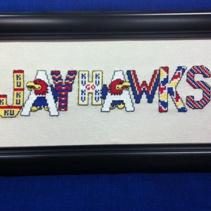 May include: A framed cross-stitch piece with the word "JAYHAWKS" in colorful letters. The letters are decorated with the Kansas University logo and the words "KU" and "GO KU".