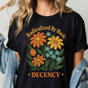 Puede incluir: Camiseta negra con un diseño floral en naranja, amarillo y azul, y el texto "Radicalized By Basic DECENCY". La camiseta tiene mangas cortas y cuello redondo.