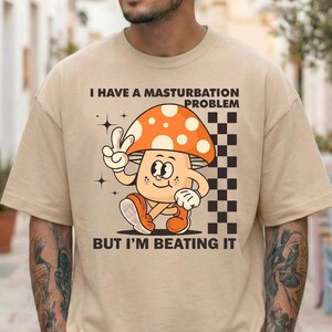 May include: Beige t-shirt with a cartoon mushroom character. The mushroom has an orange and white spotted cap, and is giving a peace sign. Text on the shirt reads "I HAVE A MASTURBATION PROBLEM BUT I'M BEATING IT".