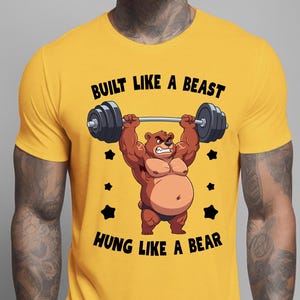 Peut inclure: T-shirt jaune moutarde avec un ours de dessin animé soulevant une barre. L'ours est brun avec un slip noir et une expression de colère. Le texte sur le t-shirt se lit "BUILT LIKE A BEAST" et "HUNG LIKE A BEAR". Des étoiles noires sont également présentes.