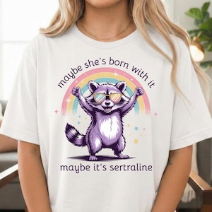 May include: White t-shirt featuring a purple raccoon wearing sunglasses, with a rainbow and the text "maybe she's born with it, maybe it's sertraline." The raccoon is standing with its arms raised.