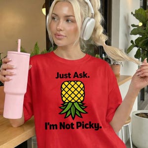 Pode incluir: Uma camiseta vermelha com o texto "Just Ask. I'm Not Picky." e um gráfico de abacaxi amarelo e verde. Um copo rosa com canudo é segurado na mão esquerda. A pessoa está usando fones de ouvido e tem cabelo loiro.