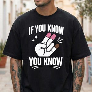 May include: Black t-shirt with the text "IF YOU KNOW YOU KNOW" above a hand gesture with pink and brown nail polish. Two small stars are on the left side of the hand gesture. The shirt is a casual style.