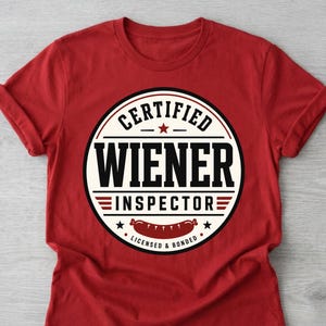 Op de afbeelding: Rood t-shirt met een cirkelvormige afbeelding met de tekst "CERTIFIED WIENER INSPECTOR" in zwart en wit. De afbeelding bevat een worstillustratie en de tekst "LICENSED & BONDED".