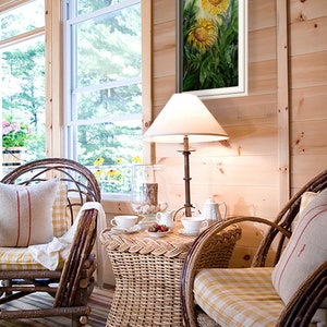 May include: A cozy interior scene featuring a framed sunflower painting, wicker furniture, and a small table set for tea. The room has wood paneling, a window, and a striped rug. A lamp with a white shade provides soft lighting.