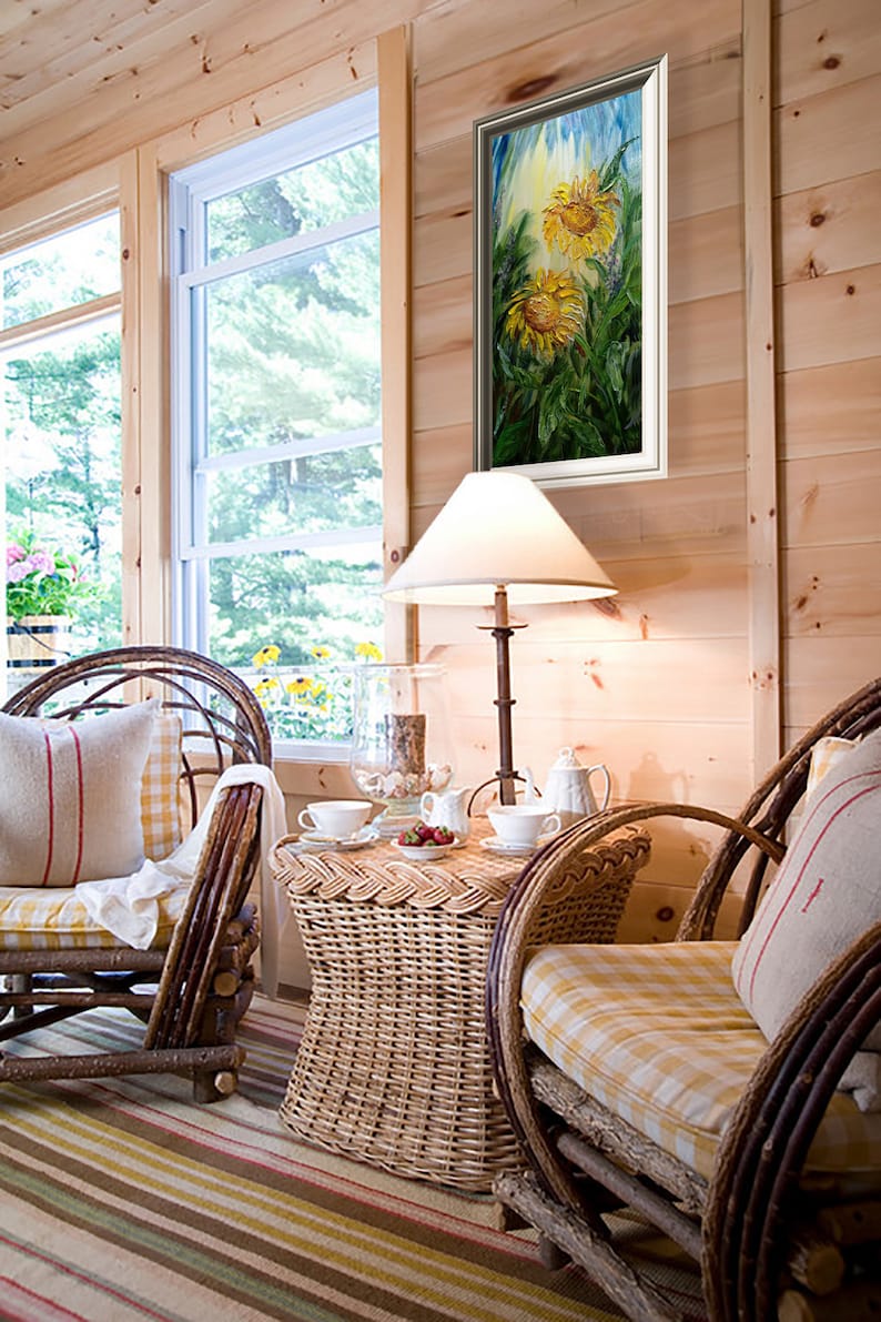 May include: A cozy interior scene featuring a framed sunflower painting, wicker furniture, and a small table set for tea. The room has wood paneling, a window, and a striped rug. A lamp with a white shade provides soft lighting.