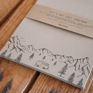 Può includere: Un blocco note di carta marrone con un'illustrazione in bianco e nero di montagne, alberi e un furgone vintage. Il blocco note ha una fascia di carta marrone con il testo "LET'S GET LOST TOGETHER" e le parole "FOR THE SCHÖNSTEN KUHLSCHRANKNACHRICHTEN, KLEINE LIEBESBRIEFE,ODER ZUR PLANUNG DEINES NÄCHSTEN ABENTEUERS" in tedesco.