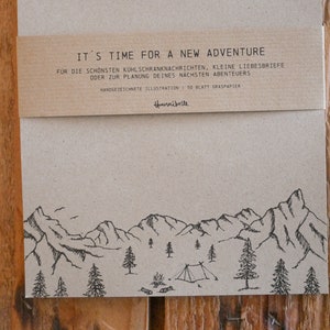 Könnte beinhalten: Ein braunes Notizbuch mit einer schwarz-weißen Illustration von Bergen und einem Campingplatz. Das Notizbuch hat eine Kopfzeile mit dem Text "It's time for a new adventure" und "Handgezeichnete Illustration 150 Blatt Graspapier".