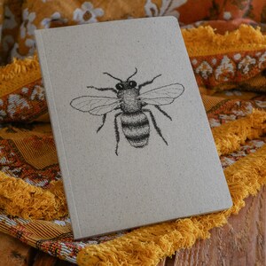 Op de afbeelding: Een grijs notitieboek met een zwart-witte tekening van een bij op de voorkant. Het notitieboek ligt op een gele en oranje gefranst stof.