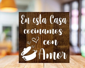 Op de afbeelding: Een vierkant houten bord met de tekst "En esta Casa cocinamos con Amor" in witte letters. Het bord heeft een witte illustratie van een kookpot en drie harten. Het hout heeft een donkerbruine vlek.