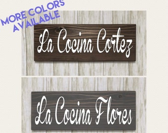 May include: Two wooden signs with white script text. The top sign is dark brown and reads "La Cocina Cortez." The bottom sign is gray and reads "La Cocina Flores." The text "MORE COLORS AVAILABLE" is in the upper left corner.