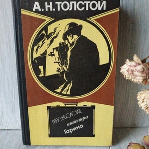 Op de afbeelding: Een vintage boekomslag met een zwart-witte illustratie van een man die door een telescoop kijkt. De titel van het boek is "A.N. TOLSTOY" en de ondertitel is "ГИПЕРБОЛОИД ИНЖЕНЕРА ГАРИНА".