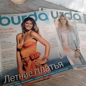 Könnte beinhalten: Zwei Ausgaben des Modemagazins "Burda" sind gestapelt. Das obere Magazin zeigt eine Frau in einem orangefarbenen Kleid, mit Text auf Russisch. Das zweite Magazin zeigt eine Frau in einer hellblauen Jacke.