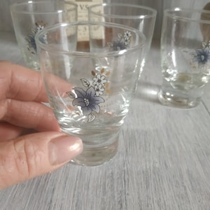 Puede incluir: Vasos de chupito transparentes con un diseño floral con flores azules y blancas con detalles dorados. Los vasos son cilíndricos con una parte superior ligeramente acampanada y una base gruesa. Se ven varios vasos.