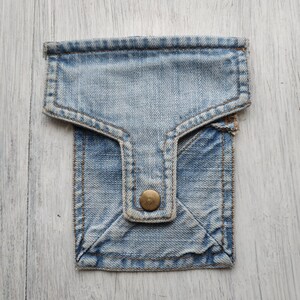 May include: A small blue denim pouch with a flap and a single button closure. The pouch is made of a soft, worn denim fabric.
