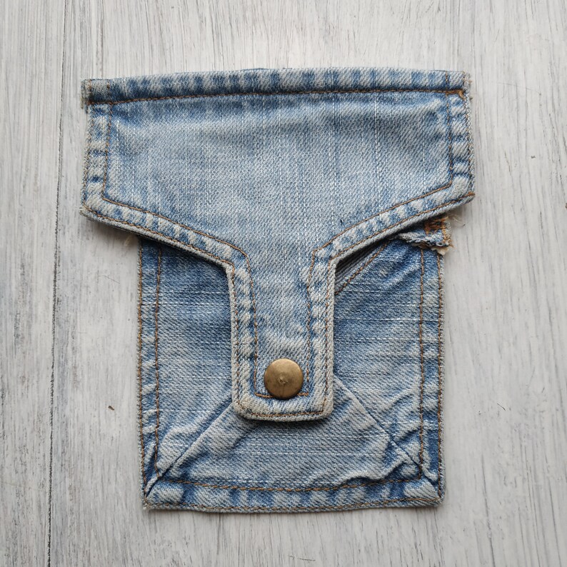 May include: A small blue denim pouch with a flap and a single button closure. The pouch is made of a soft, worn denim fabric.