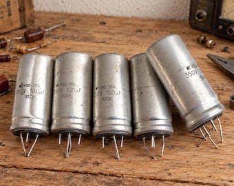 Könnte beinhalten: Fünf zylindrische, silberfarbene Elektrolytkondensatoren mit schwarzem Boden und Metallstiften. Jede Einheit ist mit dem Text K50-356, 350V, 100µF und 9308 bedruckt. Die Bauteile sind in einer Reihe angeordnet, wobei eines nach rechts geneigt ist.