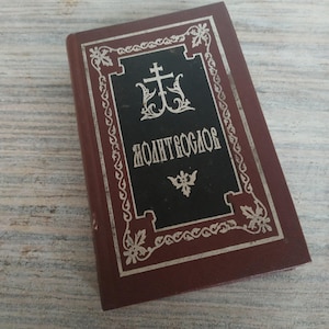 May include: A small, maroon-colored book with a black cover featuring ornate silver lettering and a cross symbol. The book's title is in a stylized font. The book is resting on a light-colored, textured surface.