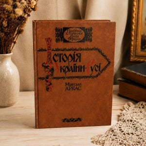 May include: A brown hardcover book titled "Istoriya Ukrayiny-Rusi" by Mykola Arkas. The cover features black decorative text and a rectangular emblem at the top with the words "Pamyatky istorychnoyi dumky Ukrayiny" printed in black ink.
