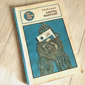 May include: A vintage book titled "AZBUKA MAKRAME" with a blue and white cover. The cover features a macrame design with scissors and a small card inside. The book is on a light wood surface.