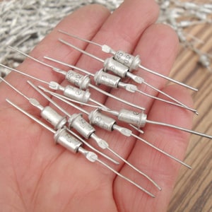 May include: A close-up of a hand holding a group of silver-colored electronic components. The components have small, cylindrical bodies with wire leads extending from each end. The components are labeled with numbers and letters, such as "145 10K" and "C8 1K7".