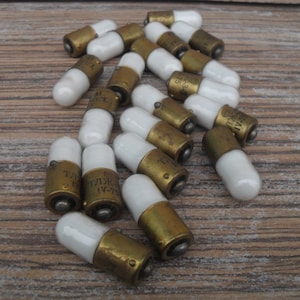 Könnte beinhalten: Eine Gruppe von 18 kleinen, weißen Glühbirnen aus Keramik mit Messingsockeln. Die Glühbirnen haben verschiedene Markierungen, darunter "TLK IV-75" und "5.5V".