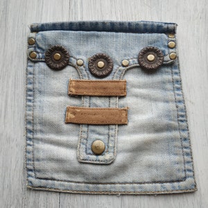 May include: A light blue denim pouch with two brown leather straps and three decorative metal accents. The pouch has a zipper closure and a small pocket on the front.